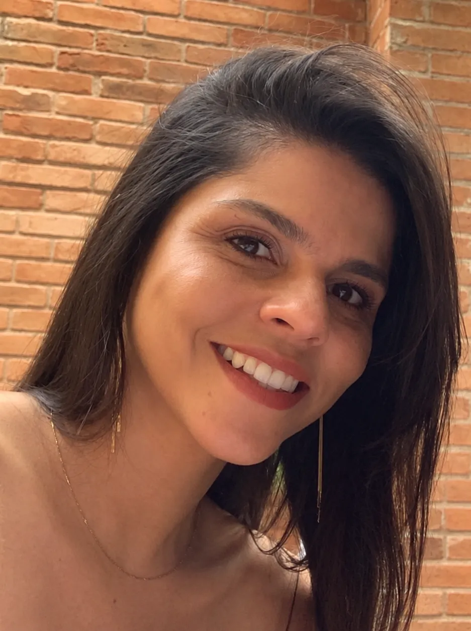 Kamilla Oliveira sorrindo, com cabelos escuros, diante de uma parede de tijolos